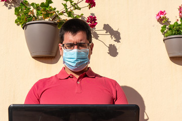 Young man with blue face mask. He is working with his laptop from the balcony of his house. He is wearing a salmon colored polo shirt and glasses. At the bottom are some hanging pots with geraniums.