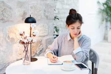 Young woman writing in notebook and reading from mobile phone in cozy cafe indoors. Freelancer...