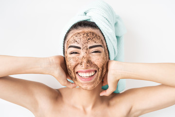 Young smiling woman applying coffee scrub mask on face - Happy girl having skin care spa day at...
