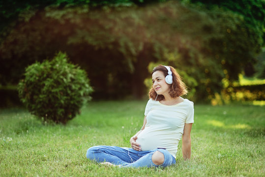 Happy young pregnant woman sitting on carpet and listening music on headphones from smartphone at park. Pregancy, Relaxation and Health care concept.