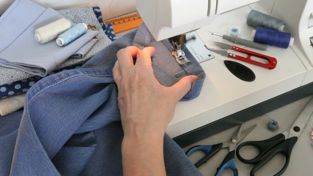 Sewing and needlework at home. Seamstress hem the bottom of the jeans on a sewing machine. Scissors, spools of thread and a bobbin on the desktop
