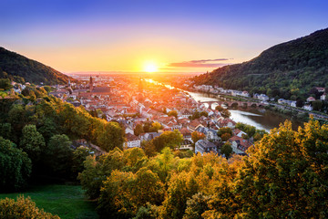 Travel shot: aerial view of Heidelberg, Germany, in beautiful sunset light, framed by colorful trees and gold and purple clear sky, with the Neckar river leading to the sun - obrazy, fototapety, plakaty