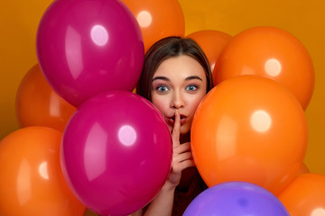 Fototapeta na wymiar Smiling caucasian girl posing with bright color air balloons showing silence gesture. Beautiful happy young woman telling secret on a birthday party. close-up
