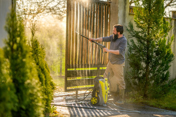 Mid adult man cleaning a wooden gate with a power washer. High water pressure cleaner used to DIY...