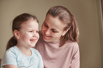 A charming little girl and her beautiful young mother use a digital tablet and smile while sitting on chairs at home