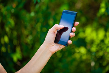 Side view of a woman's hand holding a modern slick smartphone while dialing with her thumb against summer green background