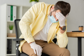 depressed businessman in latex gloves and medical mask holding hand on forehead while sitting in office