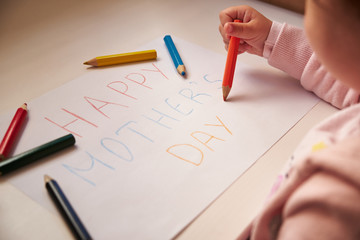 Little girl writing Happy Mothers Day on white paper colour pencil