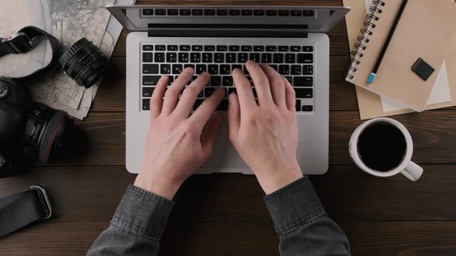 Men's hands are typing on a laptop keyboard and holding a cup of coffee. Photographer workplace, wooden table, top view
