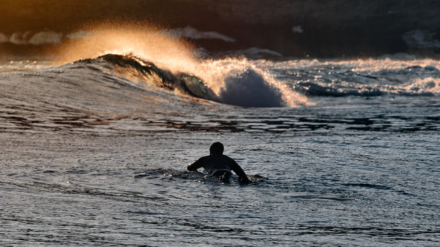 Surfer Male Silhouette Paddling Towards To Ocean Waves At Sunset.