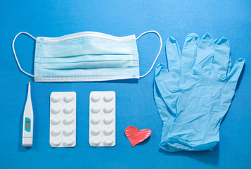 Top view of parallel COVID-19 objects  composition on a blue background showing blue surgical mask and gloves, a digital thermometer and paracetamol tablets with a red heart