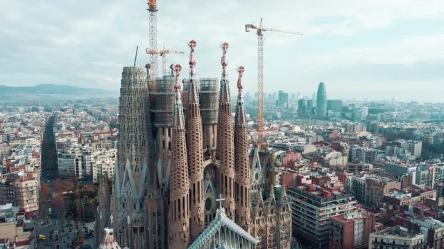 Aerial; drone view of main Barcelona attraction; majestic building towering over the rooftops of Eixample; sharp domes and unusual forms of great unfinished work of neo-gothic art; camera moves around