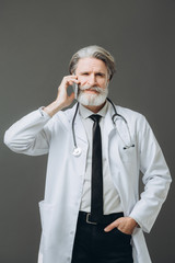 Portrait of doctor sensor on gray background. Gray-haired senor doctor dressed in white coat talking on the phone.