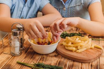 Kids soak french fries in ketchup on wooden table background. Boy and girl enjoying french fries at home during quarantine