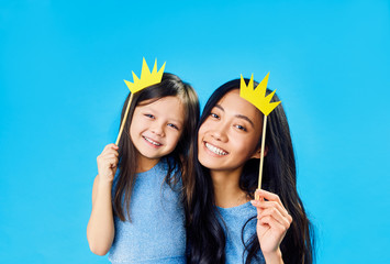 Mother and cute little daughter with a paper crown on stick on blue background have fun together