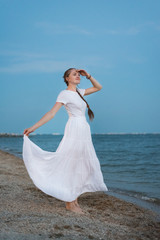 Fototapeta na wymiar Young woman in long white dress stands on shore of sea background. Vertical frame