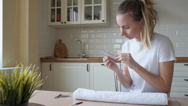 Attractive young woman doing manicure at home, smiling girl filing shaping nails with emery board