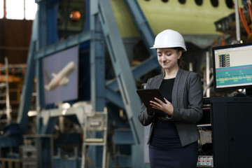 Portrait of a female factory manager in a white hard hat and business suit. Controlling the work process at the airplane manufacturer.