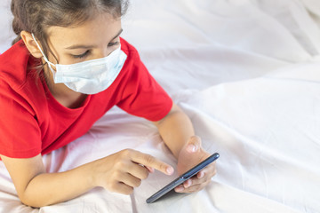 Distance schooling, learning online education. Girl wearing mask, using mobile for school or games. Laying on bed with mobile.