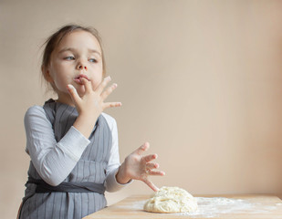 A little girl dressed as a cook stands at the table and licks the dough off her fingers. On a wooden table lay the dough in the flour