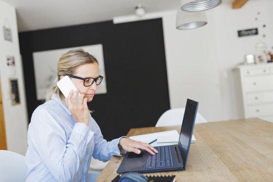 Pretty Young Woman With Blue Shirt And Glasses Doing Home Office Sitting At The Kitchen Table In Front Of Her Laptop