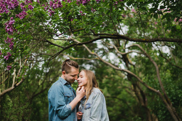 Fototapeta premium Romantic and happy caucasian couple in casual clothes hugging on the background of beautiful blooming lilac. Love, relationships, romance, happiness concept. Man and woman walking outdoors together.