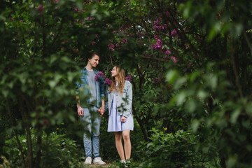 Fototapeta premium Romantic and happy caucasian couple in casual clothes hugging on the background of beautiful blooming lilac. Love, relationships, romance, happiness concept. Man and woman walking outdoors together.