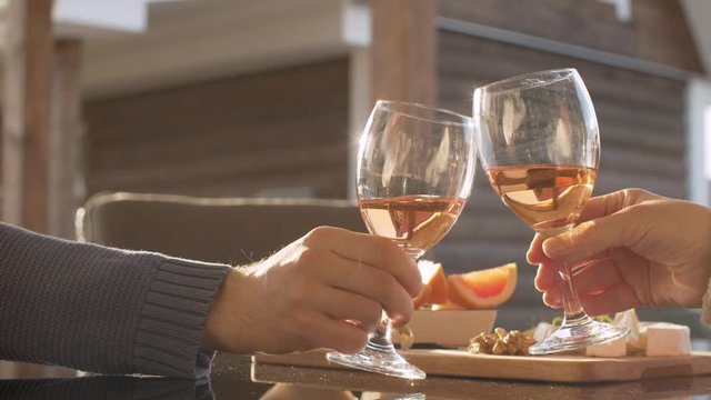 Close up hands of two people clinks wine glasses. Cheese, fruits and nuts on the table outdoors. Spring, the evening sun beautifully shining on the glass.