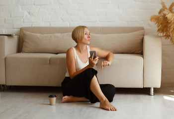 Obraz premium Young beautiful woman sitting on floor near cosy sofa with cup of coffee or tea searching in mobile