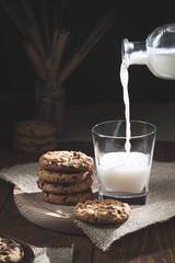 Chocolate chip cookies and milk bottle spilling milk in a glass on a wooden base, dark background. Sweet food concept.