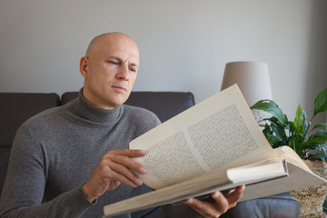 A man reads a book with interest while sitting at home on the couch.