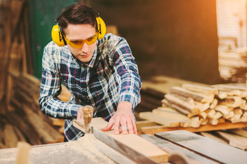 Professional carpenter in protective glasses and headphones using electric circular saw at woodworking workshop. Skilled cabinet maker working with sawing machine at sawmill. Wood production craftsman