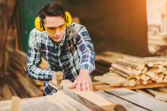 Professional carpenter in protective glasses and headphones using electric circular saw at woodworking workshop. Skilled cabinet maker working with sawing machine at sawmill. Wood production craftsman