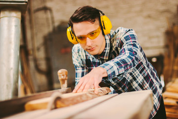 Focused professional cabinet maker in protective headphones and glasses working with sawing machine at woodworking workshop. Male carpenter using electric circular saw at sawmill. Skilled craftsman