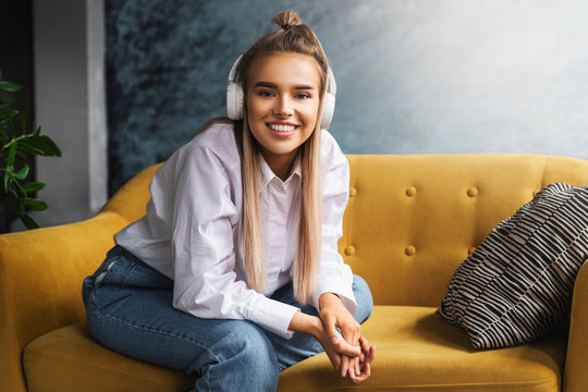 Beautiful blonde woman in white headphones sits on yellow sofa, enjoys listening to podcast. Relaxing girl in shirt