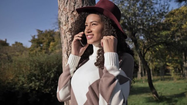 Young cheery positive african girl in hat walking outdoors in green nature park talking by mobile phone
