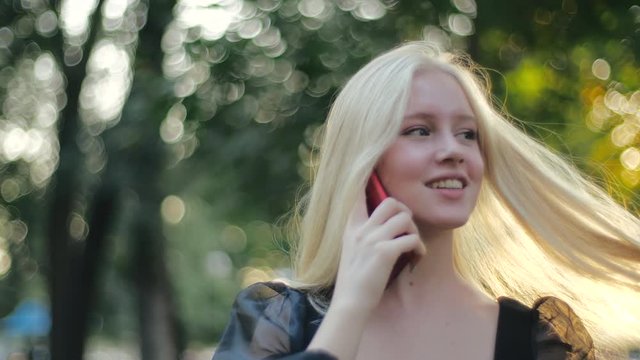 Beautiful caucasian girl fashion model walks in nature and speaks on the phone, smiles and straightens her long blonde hair. Young stylish quarantined woman outdoors in sun. Aesthetic people concept