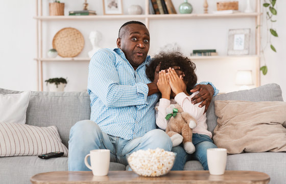 Scary movie night. Senior African American man with his little granddaughter closing face in horror at home