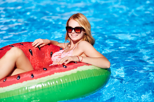Young blonde girl having fun and laughing on an inflatable watermelon pool swim ring in bikini. Attractive woman in swimwear lies in the sun on tropical vacation. Pretty female sunbathing at resort.