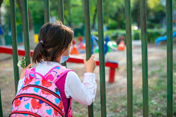 Little school girl sitting next to school fence waiting for going back to clases after pandemic outbreak