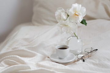 A cup of espresso coffee in bed with book, glasses and vase of white roses