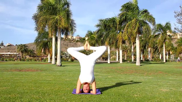 Yoga practitioner doing baddha konasana in sirsasana on sunny morning outdoors. Female yogi performing headstand bending legs with feet together at Maspalomas green park. Wellness, self care concepts