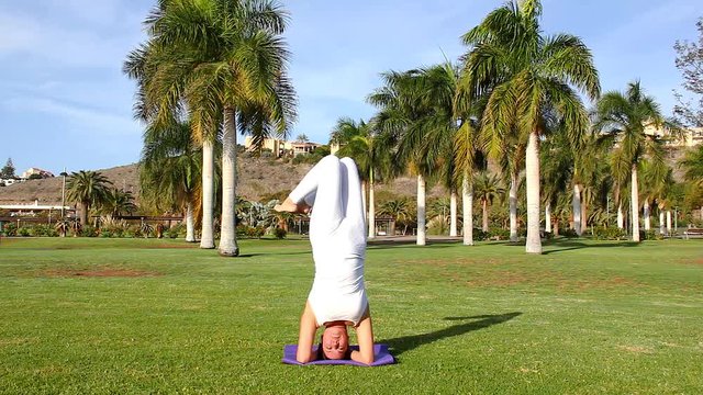 Flexible woman in parsva virasana sirsasana on purple mat at natural environment outdoors. Female yogi doing headstand bending knees and twisting body at green park. Balance, equilibrium concepts