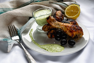 Toasted chicken drumsticks, with a brown Golden crust with fresh sauce with herbs decorated with lemon and black olives on a platter with a white textured napkin, towel