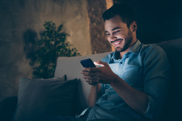 Close-up portrait of his he nice attractive cheerful cheery glad brunet guy sitting on divan using cell browsing web service at modern loft industrial style interior dark room apartment flat