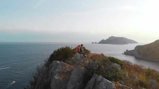 A couple of tourists are sitting on the edge of a cliff overlooking the sea. Edge of the world. Peak. The end of hiking. Destination. and an active lifestyle. Rotation. Capri island and boats distance