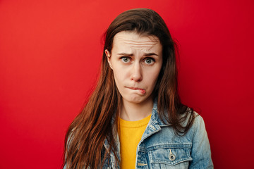 Studio shot of upset young woman purses lower lip, pouting and has displeased look, looks sadly at camera, wears denim jacket, poses against red background. People, negative face expressions concept