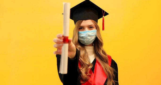 beautiful female graduate, wearing a medical protective mask, with a diploma on an isolated yellow background. Graduation, quarantine, coronavirus