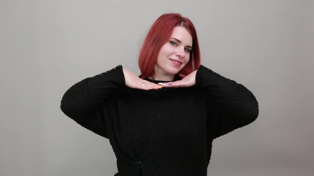 Young redhead fat lady in black sweater on grey background, cheerful woman holds her hands under head and dances with palms like in an Indian dance