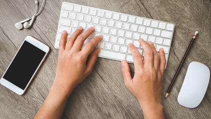 Hands holding white keyboard computer with smartphone blank screen over wood office desk table and pencil, headphones on wooden desk. Top view, flat lay.